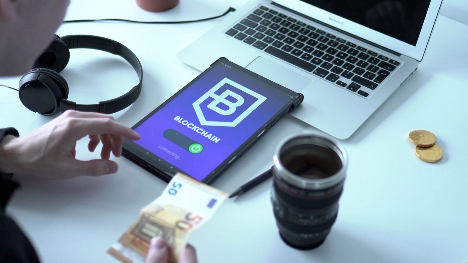 Tablet exibindo uma transação de moeda digital baseada em blockchain, ao lado de um laptop e cédulas de euro.