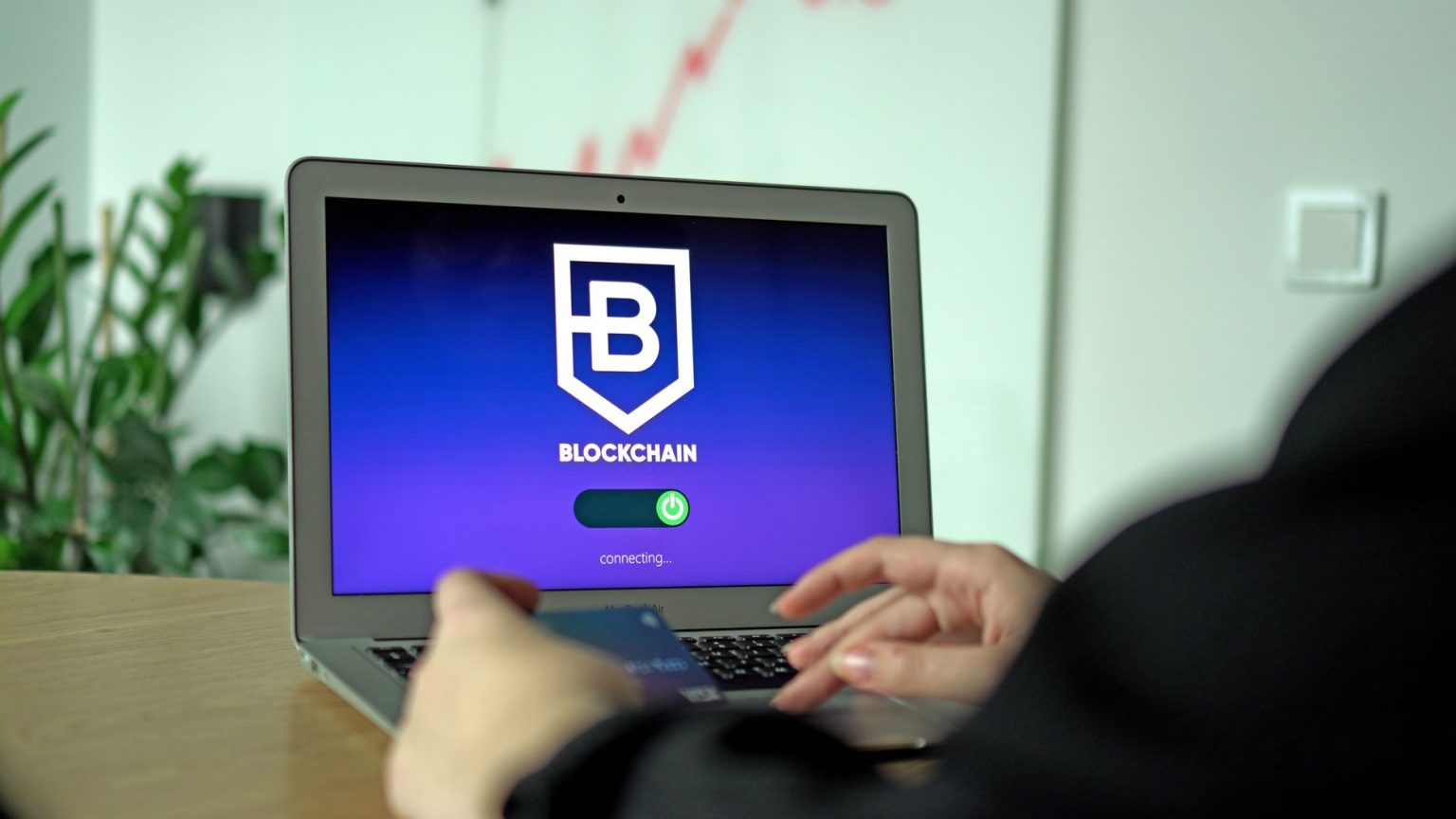 Mão segurando um cartão diante de um notebook com uma interface de conexão blockchain na tela