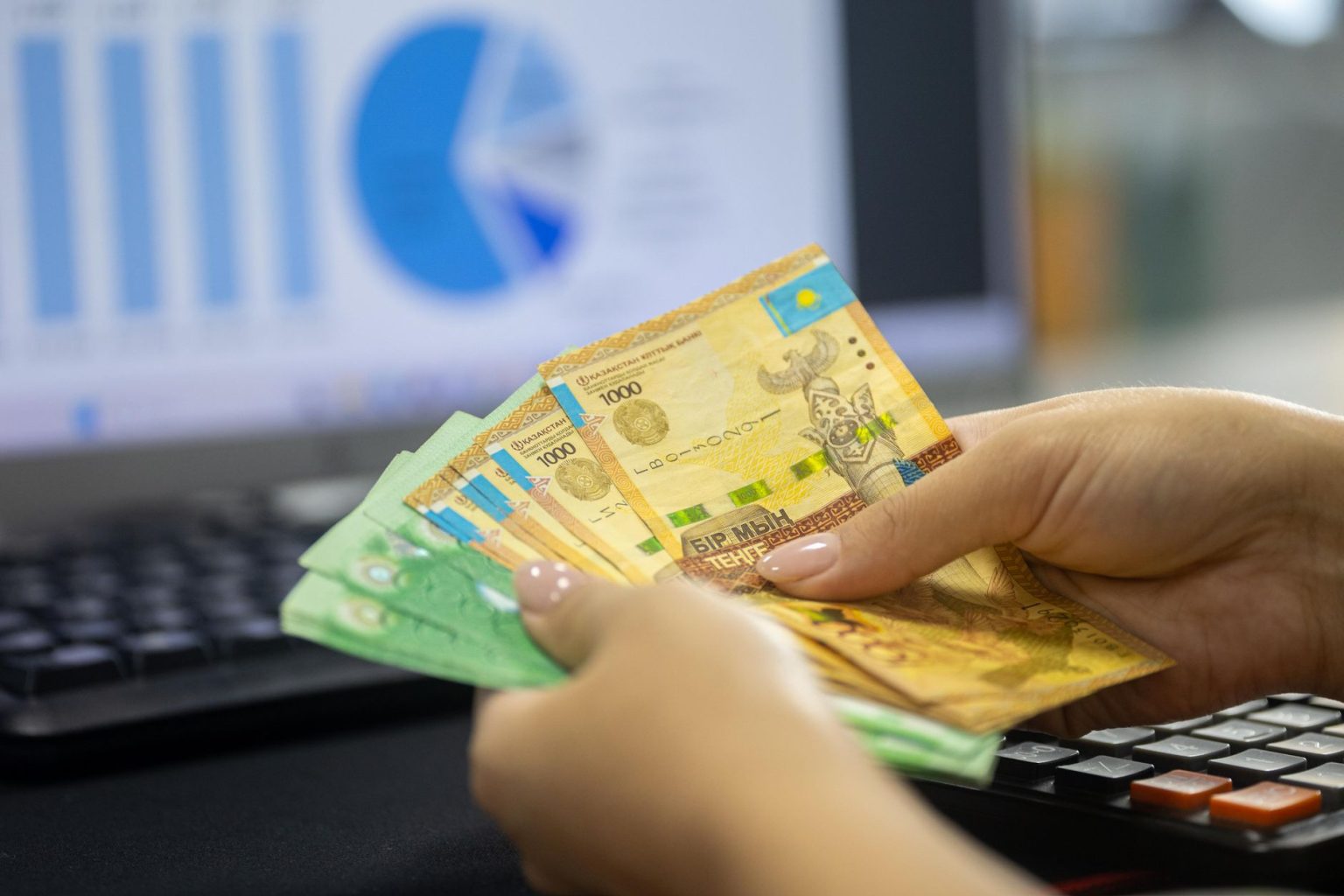 Mãos contando notas de tenge ao lado de um laptop com gráficos financeiros.
