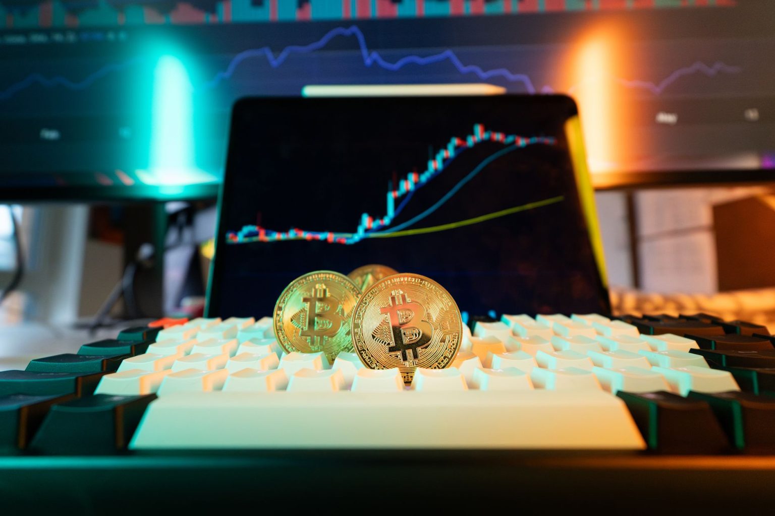 Moedas de Bitcoin sobre um teclado, com um gráfico de negociação ao fundo.