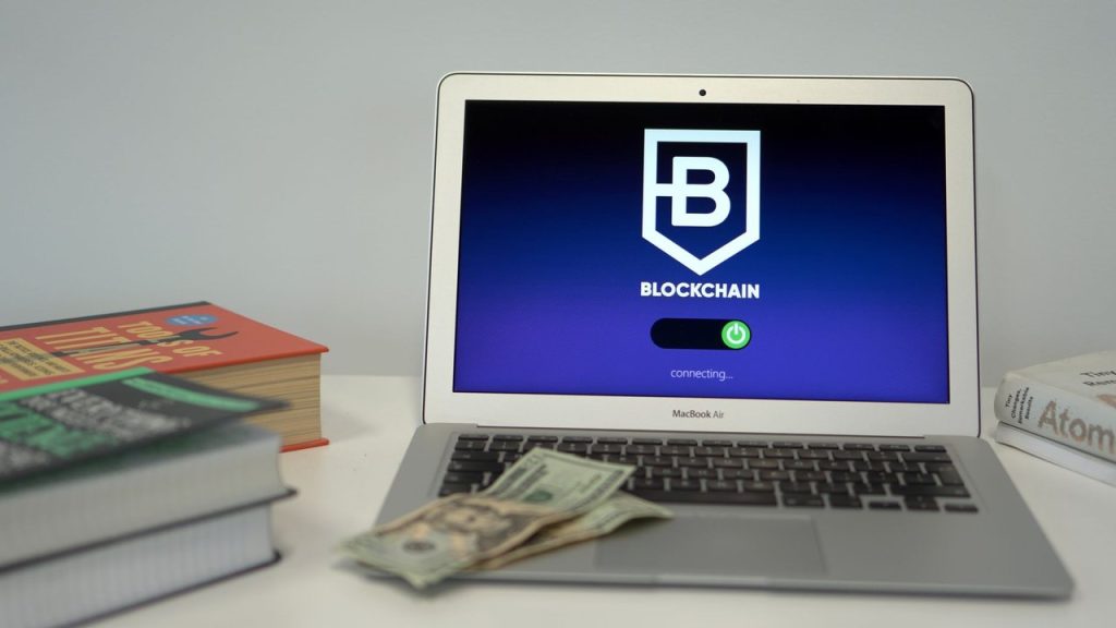 Laptop exibindo uma interface de conexão com blockchain sobre uma mesa, ao lado de livros e cédulas de dólar.