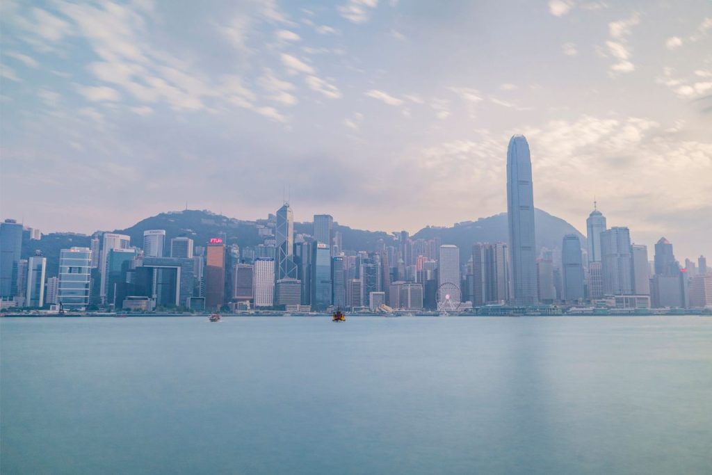 O desafio de Hong Kong para conectar stablecoins, ouro e finanças tradicionais Vista diurna do skyline de Hong Kong a partir do Victoria Harbour, com arranha-céus e área portuária.