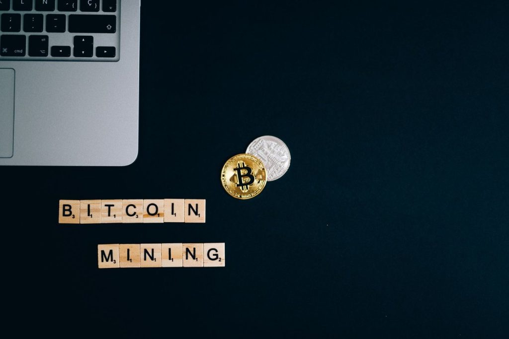 Laptop ao lado de moedas de Bitcoin e blocos com a inscrição “Bitcoin Mining” sobre uma superfície escura.